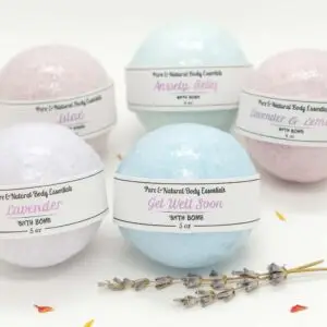 Pure & natural body essentials bath bombs in assorted pastel colors, including lavender, lilac, anxiety relief, get well soon, and lavender & lemon, arranged with a geode and lavender sprigs on a white background.
