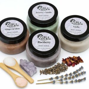 Four pure & natural body essentials body scrub jars in blackberry, ginger & clove, jasmine & vanilla, and vanilla, arranged with wooden salt spoons, amethyst crystal, dried lavender, and rose petals on a white background.