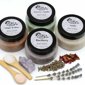 Four pure & natural body essentials body scrub jars in blackberry, ginger & clove, jasmine & vanilla, and vanilla, arranged with wooden salt spoons, amethyst crystal, dried lavender, and rose petals on a white background.