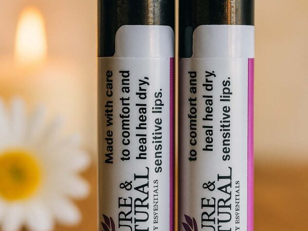 Two Pure & Natural Body Essentials lip balm tubes standing on a wooden table in warm natural light, showcasing their minimalist labels that read “Made with care to comfort and heal dry, sensitive lips.”