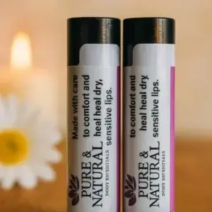 Two Pure & Natural Body Essentials lip balm tubes standing on a wooden table in warm natural light, showcasing their minimalist labels that read “Made with care to comfort and heal dry, sensitive lips.”