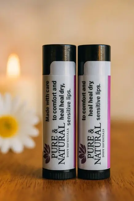 Two Pure & Natural Body Essentials lip balm tubes standing on a wooden table in warm natural light, showcasing their minimalist labels that read “Made with care to comfort and heal dry, sensitive lips.”