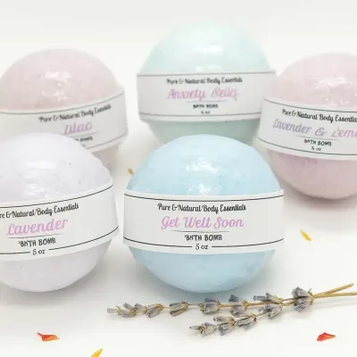 Pure & natural body essentials bath bombs in assorted pastel colors, including lavender, lilac, anxiety relief, get well soon, and lavender & lemon, arranged with a geode and lavender sprigs on a white background.