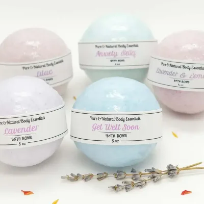 Pure & Natural Body Essentials bath bombs in assorted pastel colors, including Lavender, Lilac, Anxiety Relief, Get Well Soon, and Lavender & Lemon, arranged with a geode and lavender sprigs on a white background.